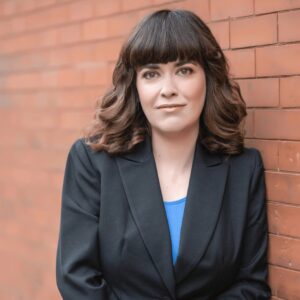 maroon brick background, white woman standing in front of it, dark blazer, blue shirt, long brown hair