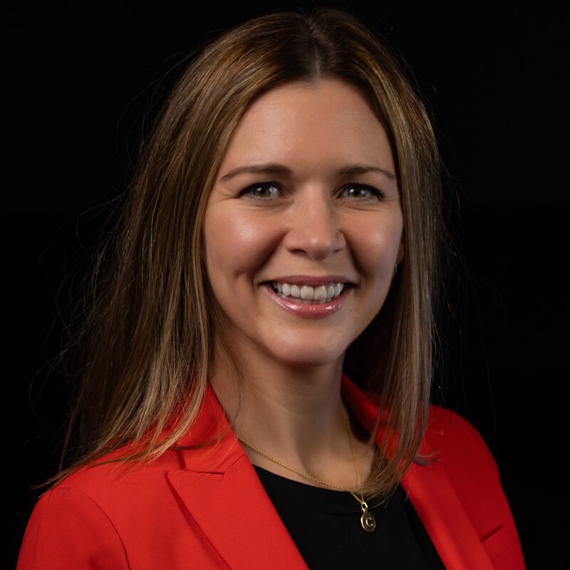 This is the headshot of an award winner smiling ahead, wearing a red blazer and black shirt in front of a black background.