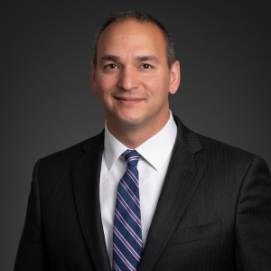 This is the headshot of an award winner smiling in a dark suit with blue striped tie in front of a black background.