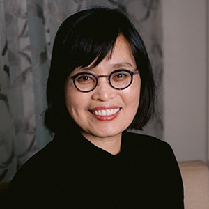 Award winner Yoon Lee smiling in front of a partially patterned grey background wearing black glasses and a black sweater