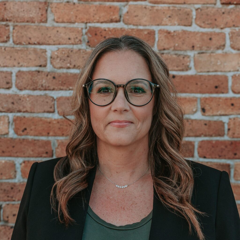 This is the headshot of an award winner standing in front of a red brick background wearing a black blazer with a green shirt and black glasses.