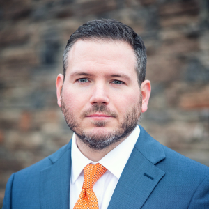 This is the headshot of an award winner looking serious in front of a green textured background in a blue suit and orange tie.